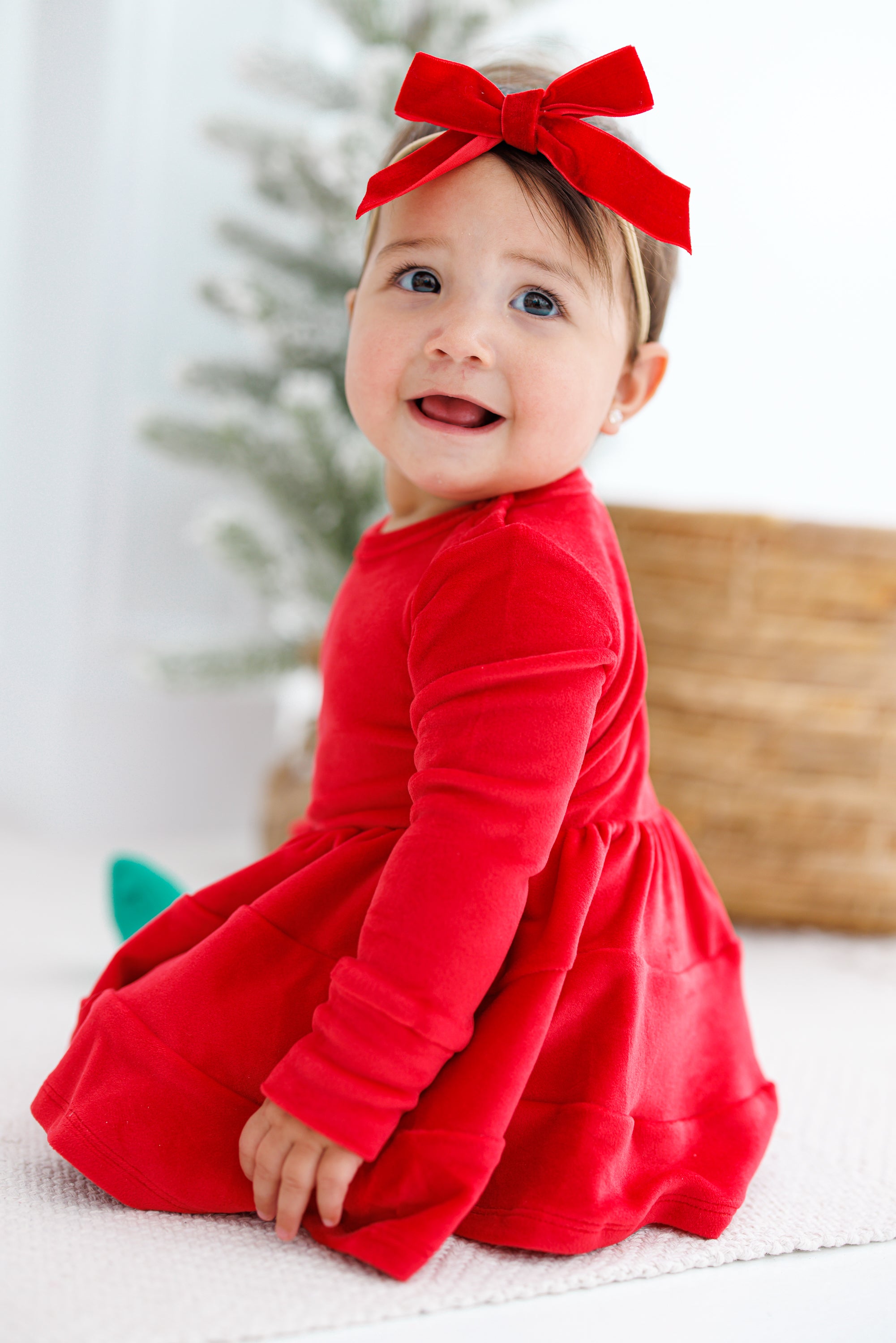 Child wearing a red velvet dress with a matching headband, sitting on a white surface.
